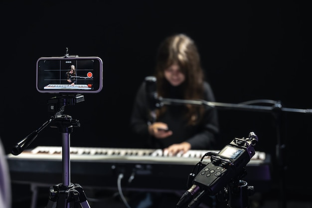 Girl musician recording video on smartphone standing on tripod, using professional microphone, blogger or music teacher shooting course in studio, sitting at the piano.