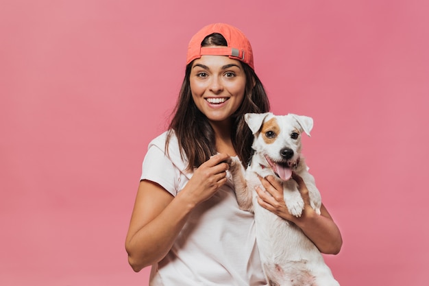 The girl in the orange baseball cap, wearing a light pink t-shirt and jeans, holds the dog by the paw sitting on her lap, smiling widely with surprised open eyes over pink wall. Happiness and dreams.