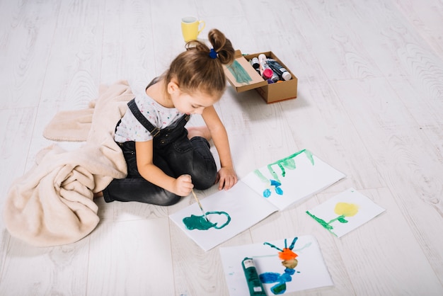 Girl painting by water colors on paper near container and sitting on floor