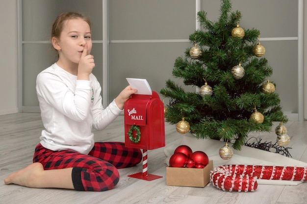 Girl in pajamas near the Christmas tree throws a letter to Santa in the mailbox Christmas gifts