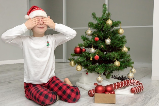 Girl in pajamas and a santa hat sits by the christmas tree with her eyes closed christmas content