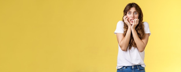 Photo girl panicking feeling scared timid insecure biting fingernails stare camera frightened clench teeth trembling fear touch face nervously feeling unconfident terrified stand yellow background