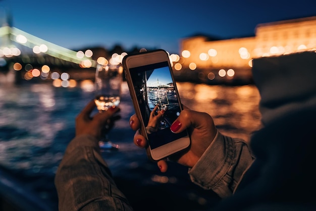 Girl photographing on phone her hand with a glass of drink on th