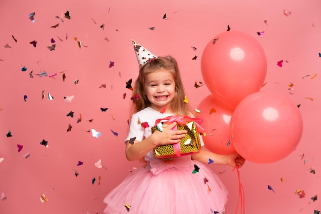 Photo a girl in a pink party hat holds a gift in front of confetti