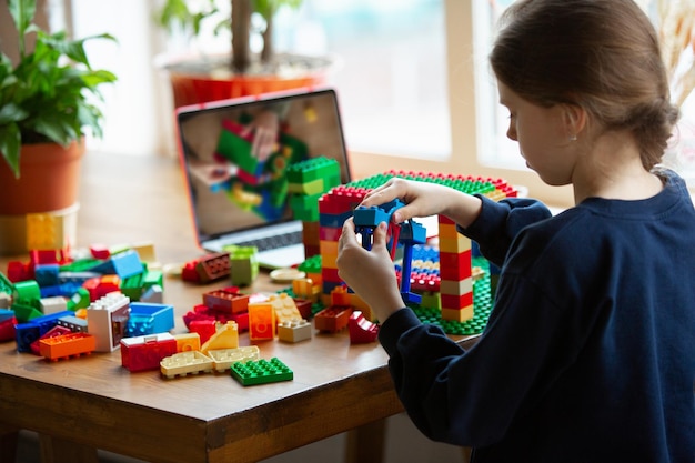 Girl playing with constructor at home, watching teacher's tutorial on laptop. Digitalization, remote education