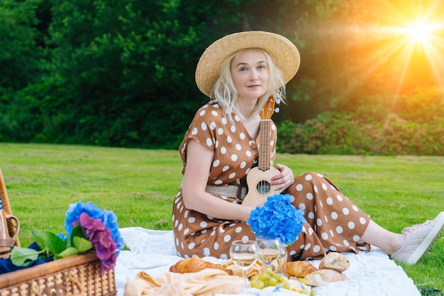 Girl in polka dot dress and hat sitting on white knit picnic blanket plays ukulele and drinking wine