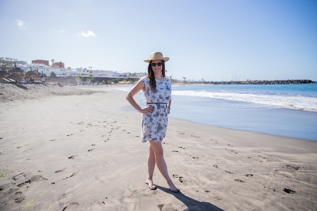 A girl poses at the beach wearing a summer hat and sunglasses, she is on a beautiful beach that looks like paradise.