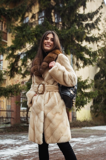 Girl posing on road on winter background Glamorous funny young woman with smile wearing stylish grey long fur coat Fur and fashion concept Beautiful people