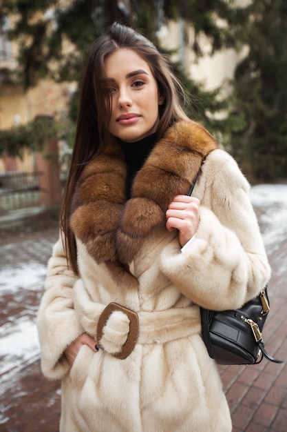 Girl posing on road on winter background Glamorous funny young woman with smile wearing stylish grey long fur coat Fur and fashion concept Beautiful people