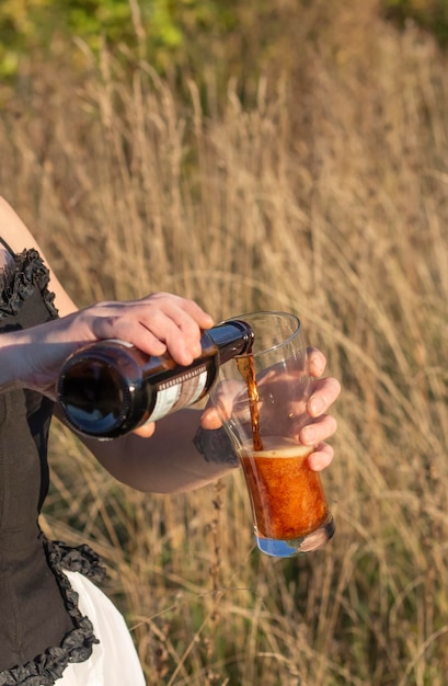 A girl pours dark beer from a bottle into a glass against the background of nature