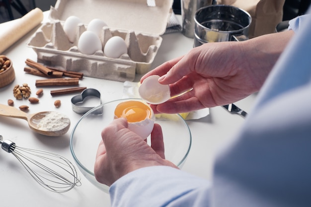 Girl preparing cookies in the kitchen, breaking an egg, close-up. A woman prepares dough for delicious pastries. Step-by-step instructions. Mixing different ingredients for the dough. Background.