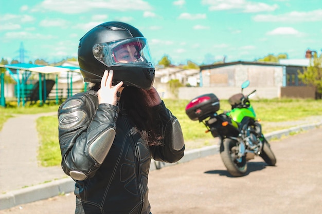 A girl in a protective motorcycle leather suit puts on a helmet against the backdrop of a motorcycle