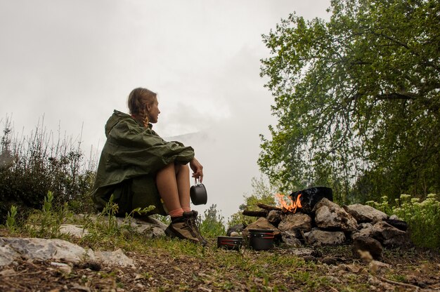 Girl in the raincoat sitting near the pot on the campfire