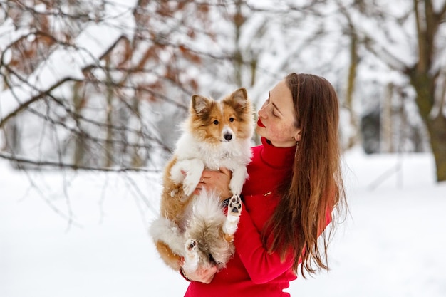 A girl in a red dress with a Sheltie dog in her arms. Winter background.