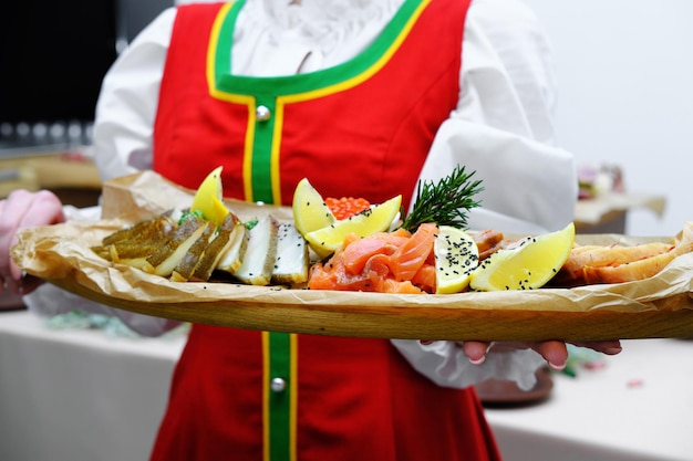 A girl in a red Russian folk dress holds a wooden tray with a fish platter in her hands Buffet in Russian style