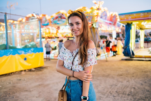 Girl in round glasses brown hair in a denim skirt and a round handbag on the background of an amusement park