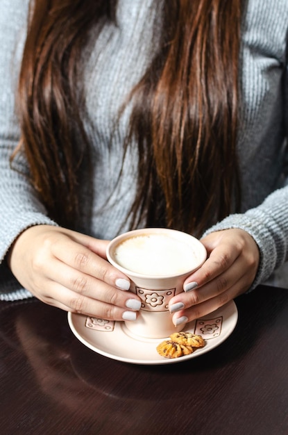 Girl's hands with cup of coffee with milk accompanied by mini cookies on wooden table