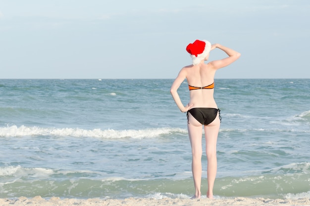 Girl in Santa hat and bathing suit looks into the distance. Sea shore.