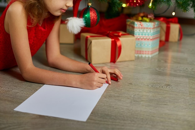 Girl in Santa hat writes letter to Santa near christmas tree at home