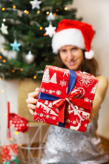 A girl in a shiny dress and a santa claus hat holds a beautifully sealed gift against the background of a decorated christmas tree preparing for the new year and christmas