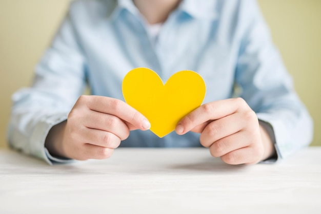 A girl in a shirt holds a yellow heart in her hands while sitting at the table Heart in hands