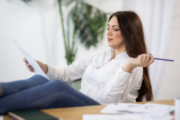 A girl in a shirt sits near the table and draws ideas on paper and in a notebook in the room