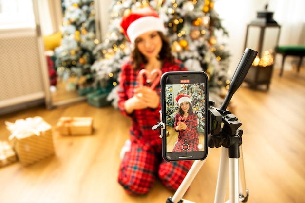 Girl shooting her christmas video blog by smartphone on tripod. She wearing red pajamas and santa claus hat.