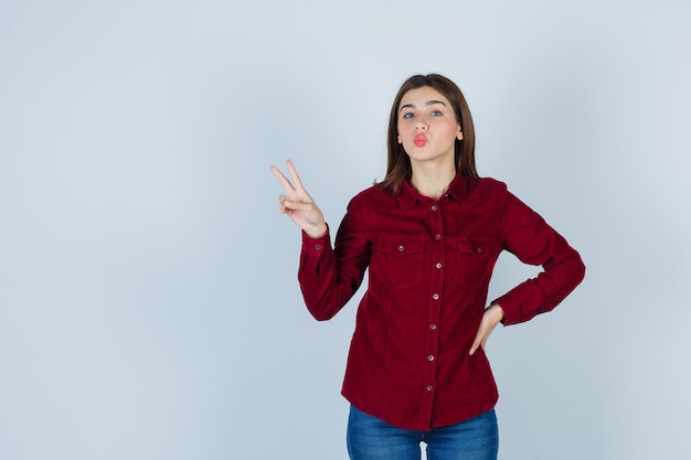 girl showing victory gesture, pouting lips in burgundy blouse and looking positive