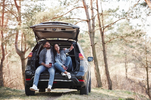 Girl shows next destination. Sitting on rear part of automobile. Enjoying the nature. Couple have arrived to the forest on their brand new black car