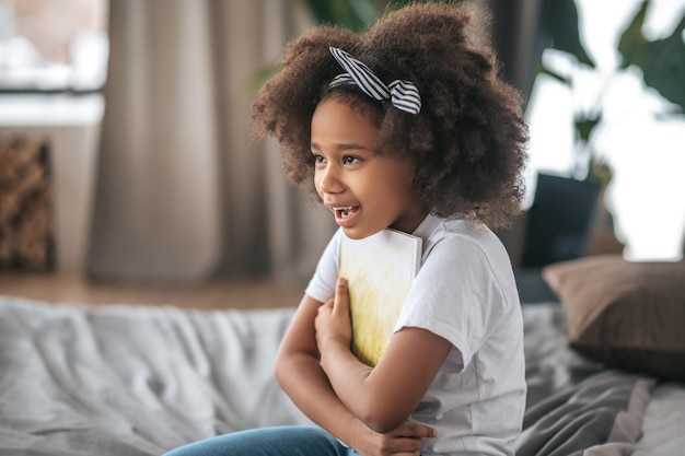 A girl sigtting on a ned with a book in hands and smiling