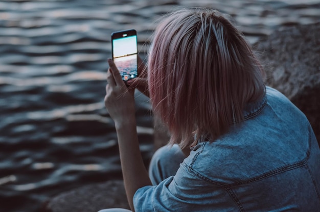 Girl sits in denim overalls and photographs the sunset through the phone