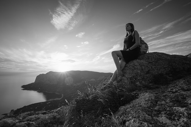 A girl sits on the edge of the cliff and looking at the sun sea and mountains. The idea and concept of recreation, freedom, summer, outdoor activities and travel