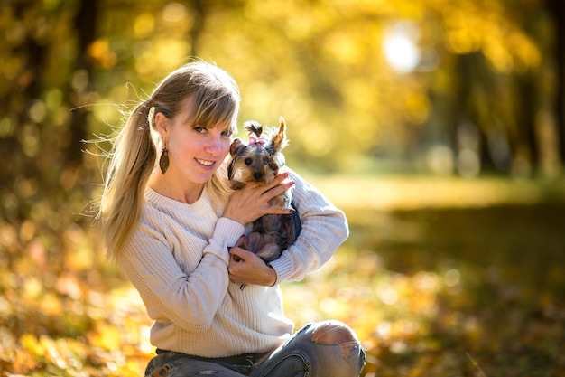 Girl sits on the ground in the autumn forest and plays with Yorkshire Terrier small dog