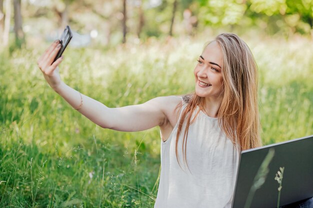 Girl sits outdoors and works at a laptop makes a selfie on the phone freelance selfeducation the concept of remote learning and outdoor work