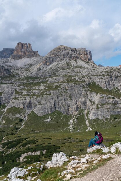 The girl sits with her back on a stone and looks at the majestic Dolomites in Italy with a view of t