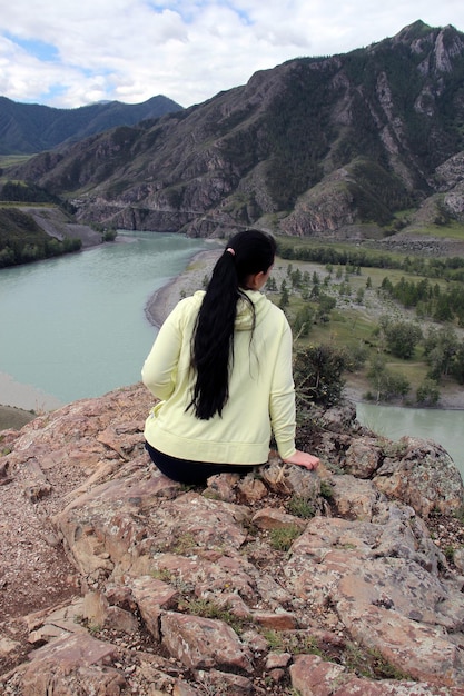 The girl sits with her back to us on the edge of the mountain Against the backdrop of a blue river mountains and forest