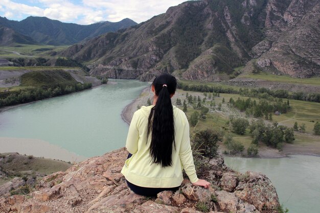 The girl sits with her back to us on the edge of the mountain Against a blue river and mountains