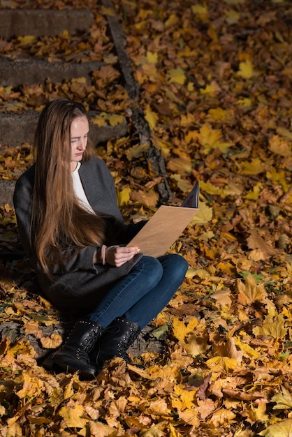 Girl sitting in autumn Park with book in hand Young woman reading book in autumn forest Vertical frame