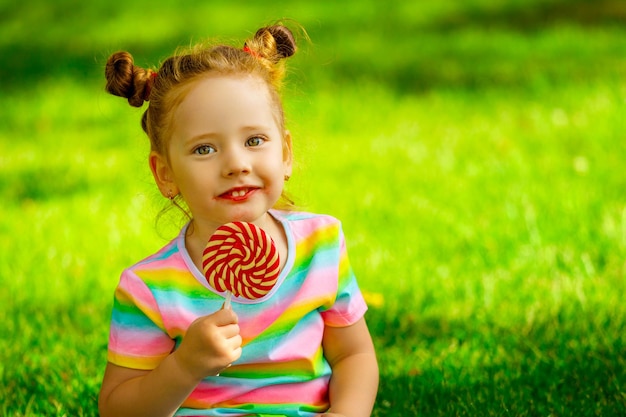 Girl sitting on the grass in the park and holding a lollipop in her hand