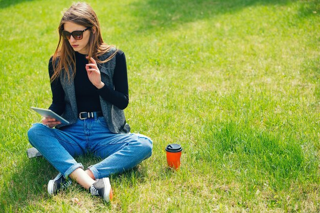 Girl sitting on the grass with coffee and tablet on the street.