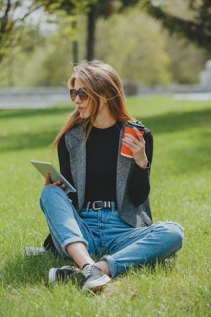 Girl sitting on the grass with coffee and tablet on the street.