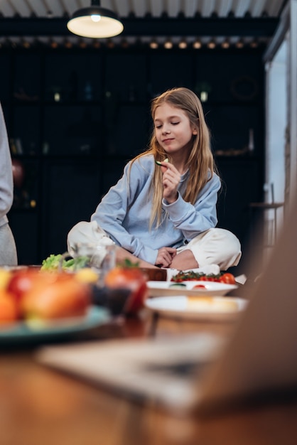 Girl sitting on the kitchen table eating a cucumber.