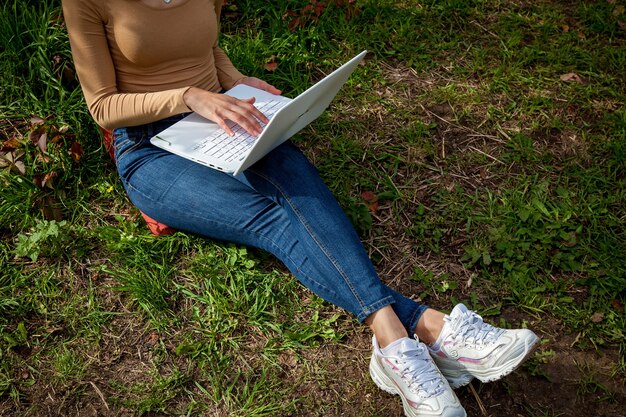 Girl sitting near a tree in the park and working on a laptop. Freelance work outdoors. Working outside the office due to the Covid 19 pandemic