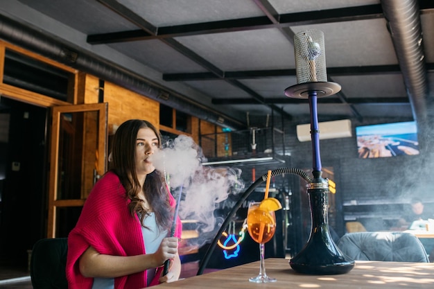 Photo girl sitting at a table in the summer smokes a hookah young beautiful girl drinks aperol hookah and aperol on the table