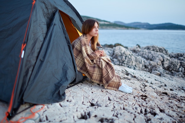 Girl sitting under tents, wrapped in plaid, holding cup of fragrant tea.