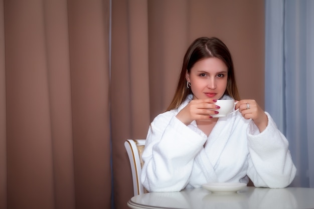 Girl of Slavic appearance in coat with white Cup is sitting at table