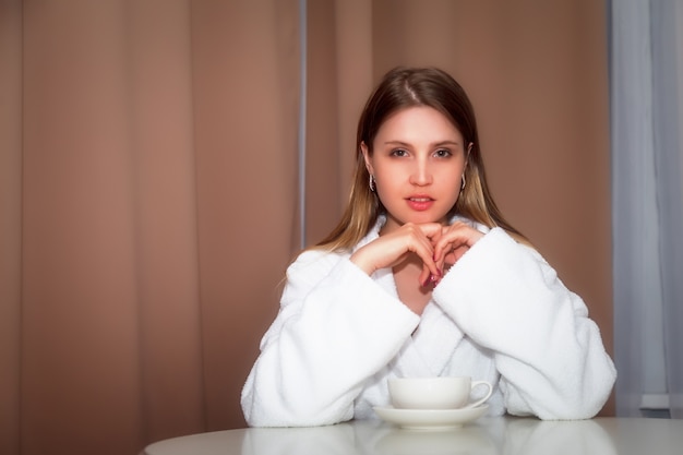 Girl of Slavic appearance in coat with white Cup is sitting at table