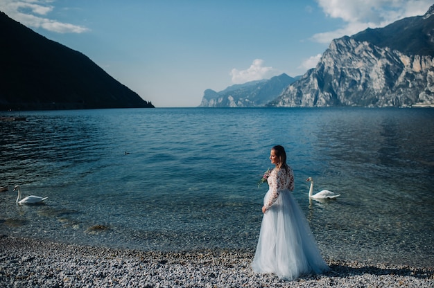 A girl in a smart white dress walks along the embankment of lake Garda.A woman is photographed against the background of a mountain and lake in Italy.Torbole