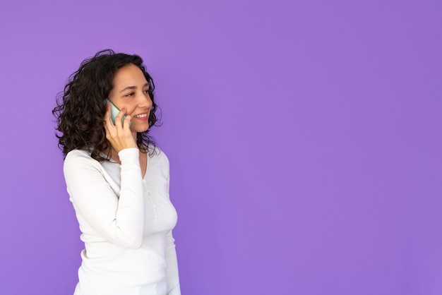 Girl smiles while talking on the phone. Purple background and copy space. White casual shirt. Curly hair.