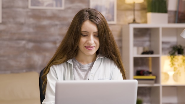 Girl smiling while working on laptop in living room. Boyfriend eating chips in the background.